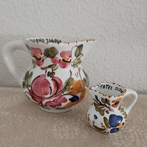 May include: Two white ceramic pitchers with hand-painted floral designs in pink, green, and orange. The larger pitcher features the text "AVIANO T.66044" along the rim, while the smaller pitcher displays the text "HOTEL OLIV" on its upper edge.