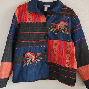 May include: A button-up jacket featuring a patchwork design of blue denim, black fabric, and orange ruffled panels. The garment includes embroidered floral motifs in orange and green, horizontal red stitching accents, a standard collar, and long sleeves.