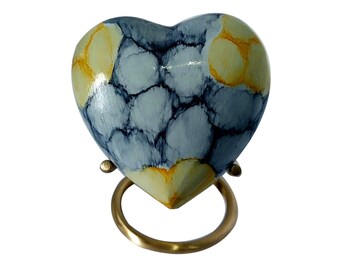 Op de afbeelding: Een kleine, hartvormige houten urn met een blauw en geel marmerpatroon. De urn staat op een gouden metalen standaard.