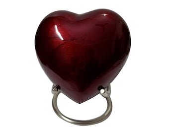 Op de afbeelding: Een kleine, hartvormige crematie urn van metaal met een glanzende, rode afwerking. De urn staat op een zilveren standaard met een ring.