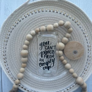May include: A clear glass cup with a black vinyl decal that reads "You can't pour from an empty cup". The cup is sitting on a white woven tray with a wooden bead garland.
