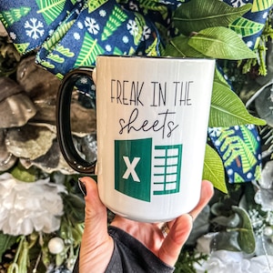 May include: White ceramic mug with a black handle and rim, featuring the text "FREAK IN THE sheets" above an Excel logo. The mug is held against a backdrop of a festive wreath with blue and green accents.