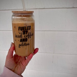 May include: A glass tumbler with a bamboo lid and a straw filled with iced coffee. The tumbler has a black graphic that reads "Fueled by iced coffee and peloton".