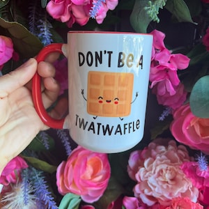 May include: White ceramic mug with a red rim and handle. The mug has a cartoon waffle with a smiling face and the text "Don't be a Twatwaffle".