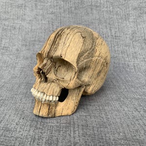 Puede incluir: Una escultura de cráneo humano tallada en madera de color tostado claro con vetas de color marrón oscuro. La pieza presenta cuencas oculares profundas, una cavidad nasal y una hilera de dientes lisos de color blanco roto en la mandíbula.
