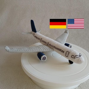 Puede incluir: Un avión de juguete de crochet con un cuerpo gris y blanco y detalles azules. El avión está sentado en un soporte para pasteles blanco. Las banderas de Alemania y los Estados Unidos están en el fondo.