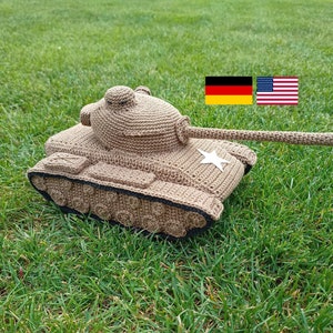 Könnte beinhalten: Ein gehäkelter Spielzeugpanzer in Braun mit einem weißen Stern an der Seite. Der Panzer steht auf grünem Gras. Im Hintergrund sind zwei Flaggen zu sehen, eine deutsche Flagge und eine amerikanische Flagge.