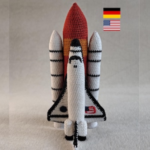 Könnte beinhalten: Gehäkeltes Raumschiff-Spielzeug in Weiß, Schwarz, Rot und Orange. Das Spielzeug zeigt die amerikanische Flagge und ist ein weiches, plüschiges Objekt. Das Shuttle hat ein detailliertes Design mit Flügeln und einer spitzen Nase.