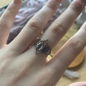Può includere: Un anello d'argento con un design delicato e intricato che presenta un motivo floreale. L'anello è indossato sul dito medio di una mano.