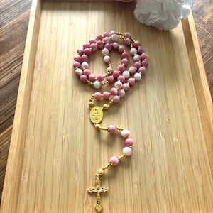 May include: A rosary featuring alternating pink and white spherical beads accented with small gold-toned spacers. The piece includes a gold-toned oval centerpiece depicting a religious figure and a gold-toned crucifix at the bottom end.