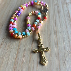Puede incluir: Un rosario de cuentas de colores con una cruz de oro. Las cuentas son de color rosa, naranja, amarillo, azul y verde.