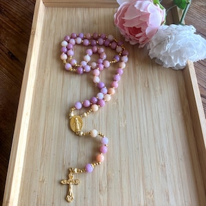 May include: A rosary with purple, pink, and peach-colored beads. The rosary has gold-colored accents and a gold-colored crucifix. The rosary is displayed on a wooden tray with pink and white flowers.