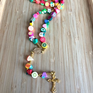 Puede incluir: Un rosario de temática frutal de colores con una cruz dorada y cuentas. Las cuentas están hechas de arcilla polimérica y presentan diversas frutas, como fresas, sandía, limones y piñas.