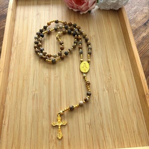 May include: A rosary featuring brown and tan stone beads and gold-coloured detailing. The rosary includes a gold-coloured crucifix and a gold-coloured medallion. It is presented on a wooden tray.