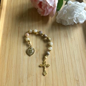 May include: A single-decade rosary featuring smooth, multi-colored round beads in shades of cream, peach, and grey, accented with gold-toned metal spacers. The rosary includes a gold-toned metal crucifix and a heart-shaped pendant with a crown detail.
