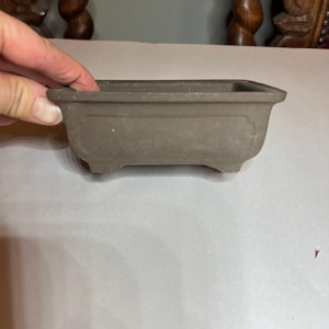 May include: A rectangular, brown bonsai pot with a slightly curved design on the sides. The pot has small feet and is made of a matte material. The pot is empty and ready for planting.