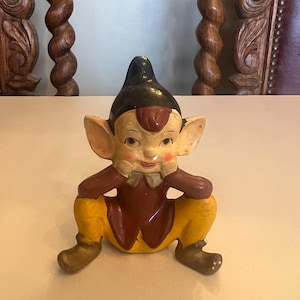 May include: A vintage ceramic elf figurine, seated with a black hat, red and brown clothing, and yellow trousers. The elf has large ears, rosy cheeks, and a small bow tie. A collectible item.