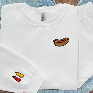 以下が含まれることがあります： 前面にホットドッグのグラフィック、袖にマスタードとケチャップのボトルがプリントされた白いスウェットシャツです。