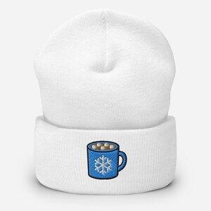 May include: White beanie with a blue embroidered mug design. The mug features a snowflake and marshmallows, suggesting a winter theme. The beanie is a classic, comfortable style.