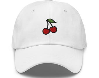May include: White baseball cap with a red and green embroidered cherry design. The cap has a curved brim and an adjustable strap at the back. The cherry design is a simple, cartoon-style illustration.
