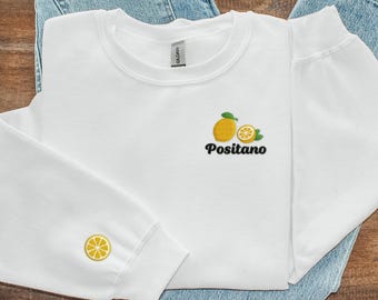 Könnte beinhalten: Weißer Pullover mit einem Zitronen- und Zitronenscheiben-Motiv und dem Text "Positano" auf der Vorderseite. Der Pullover hat ein Zitronenmotiv auf dem linken Ärmel.