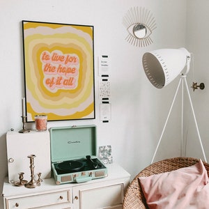 May include: A retro-style record player in a light green colour with a black record on the turntable. The record player is sitting on a white dresser with three drawers. There is a framed print with the text "to live for the hope of it all" in a yellow, orange, and pink colour scheme. The print is hanging on a white wall. There is a wicker chair with a pink cushion in the corner of the room.