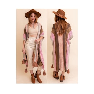 May include: A long, striped knit cardigan with tassels. The cardigan is in shades of brown, pink, and white. The model is wearing the cardigan with a white tank top, white pants, and brown boots.