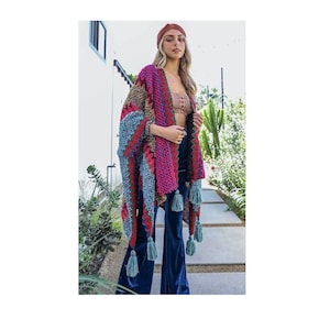 May include: A colorful, open-front knit shawl with a geometric pattern in shades of pink, blue, and brown. The shawl features long tassels in a matching blue-green color. The model is wearing the shawl with dark blue velvet pants.