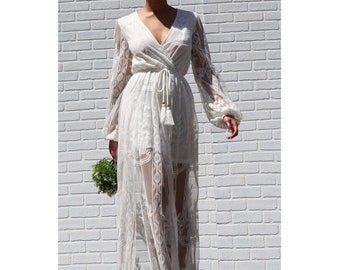 May include: A white lace maxi dress with long sleeves and a V-neckline. The dress has a tie at the waist and a flowing skirt.