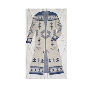 Op de afbeelding: Lange, openvallende cardigan in neutraal beige met een geometrisch patroon in blauwtinten. De cardigan heeft lange mouwen, zakken en een contrasterende blauwe bies langs de randen en manchetten. Het ontwerp bevat driehoeken en ruiten.