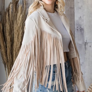 May include: Beige suede jacket with long fringe detailing. The jacket has a classic collar and long sleeves. The model is wearing denim shorts and a white top. The jacket is a neutral color and can be worn with many outfits.