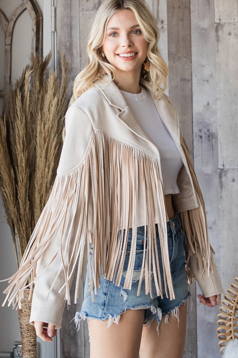 May include: Beige suede jacket with long fringe detailing. The jacket has a classic collar and long sleeves. The model is wearing denim shorts and a white top. The jacket is a neutral color and can be worn with many outfits.