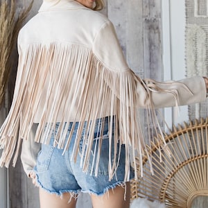 May include: A cream-colored suede jacket with long fringe detailing across the back and sleeves. The jacket is paired with denim shorts. The overall style is bohemian and western-inspired.