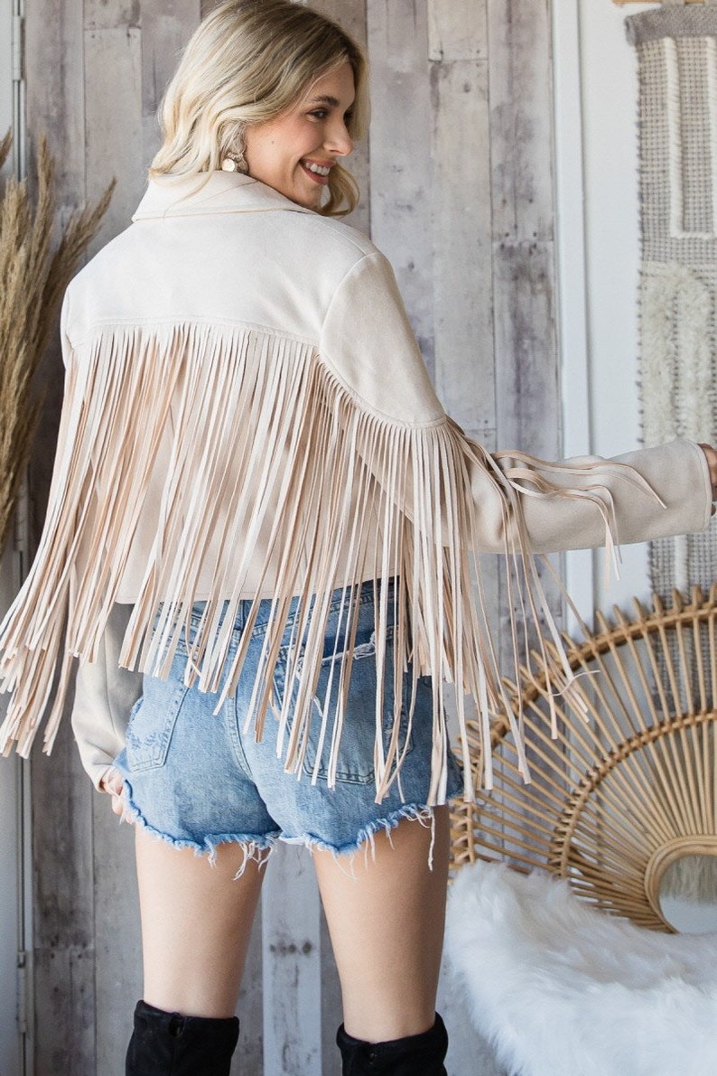 May include: A cream-colored suede jacket with long fringe detailing across the back and sleeves. The jacket is paired with denim shorts. The overall style is bohemian and western-inspired.