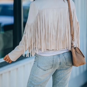 May include: A beige jacket with long fringe detailing across the back and shoulders. The jacket has a collared neckline and long sleeves. The person is wearing light wash blue jeans and a brown purse with a shoulder strap.