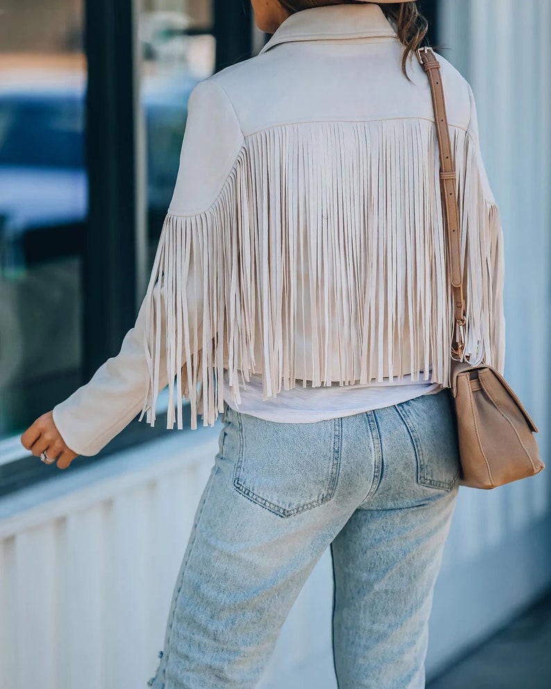 May include: A beige jacket with long fringe detailing across the back and shoulders. The jacket has a collared neckline and long sleeves. The person is wearing light wash blue jeans and a brown purse with a shoulder strap.