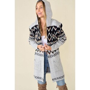 May include: A long, gray hooded cardigan with a black and white geometric pattern on the upper chest and sleeves. The cardigan has two front pockets and a repeating pattern at the hem. The model is wearing blue jeans and brown boots.