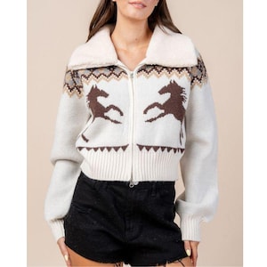 May include: Cream-coloured zip-up sweater with brown horse silhouettes and a faux fur collar. The sweater features a geometric pattern at the top and bottom, with long sleeves and a ribbed hem. A stylish and cosy outerwear option.