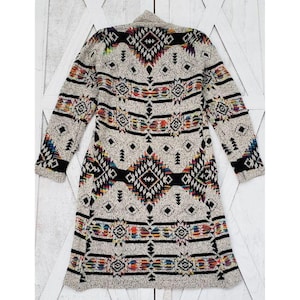 May include: A long, open-front cardigan in a neutral color with a geometric pattern in black, red, yellow, and blue. The sweater features long sleeves and a shawl collar. The design incorporates diamond and stripe motifs.
