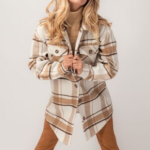 Puede incluir: Una camisa-chaqueta a cuadros beige y blanca con dos bolsillos delanteros. La chaqueta se combina con pantalones acampanados marrones y un sombrero fedora beige. Se ve un collar dorado. El conjunto es informal y elegante.