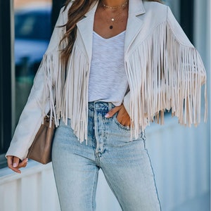 May include: Cream-colored fringed jacket with a notched collar and long sleeves. The jacket is paired with a white V-neck top and light-wash blue jeans. A small brown purse is carried on the arm. The outfit is completed with layered necklaces.