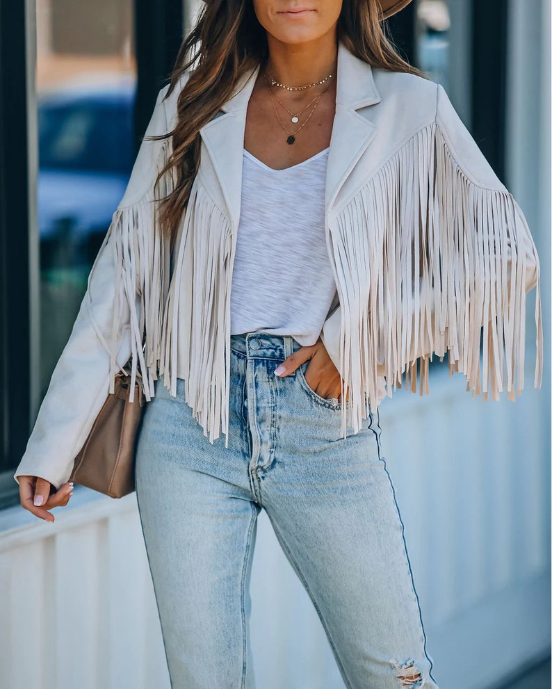 May include: Cream-colored fringed jacket with a notched collar and long sleeves. The jacket is paired with a white V-neck top and light-wash blue jeans. A small brown purse is carried on the arm. The outfit is completed with layered necklaces.
