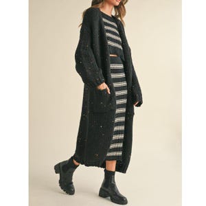 May include: A black knit cardigan with colorful flecks, worn over a matching striped crop top and skirt. The long cardigan has pockets and is paired with black ankle boots. The outfit is a stylish ensemble.