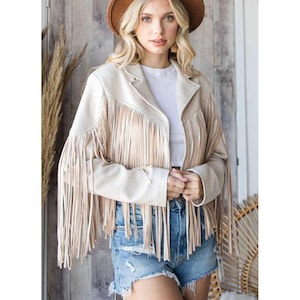 May include: Beige suede jacket with long fringe detailing on the front and sleeves. The jacket has a classic collar and a tailored fit. The model is wearing a brown hat and denim shorts. The jacket is a stylish outerwear piece.