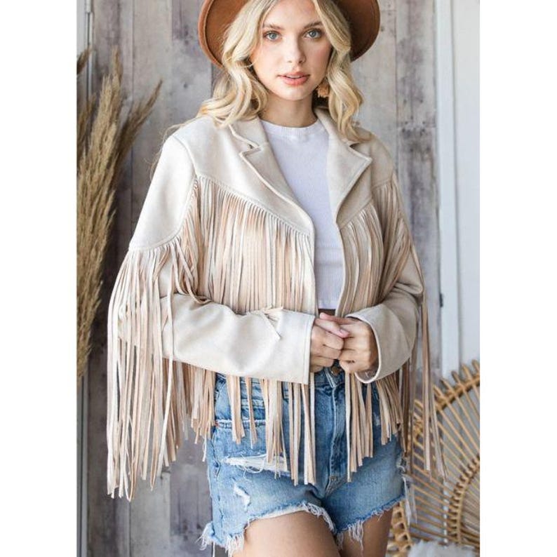 May include: Beige suede jacket with long fringe detailing on the front and sleeves. The jacket has a classic collar and a tailored fit. The model is wearing a brown hat and denim shorts. The jacket is a stylish outerwear piece.