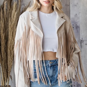 May include: Beige suede jacket with long fringe detailing. The jacket has a classic collar and is worn over a white crop top and denim shorts. The jacket's fringe adds a bohemian flair to the outfit.