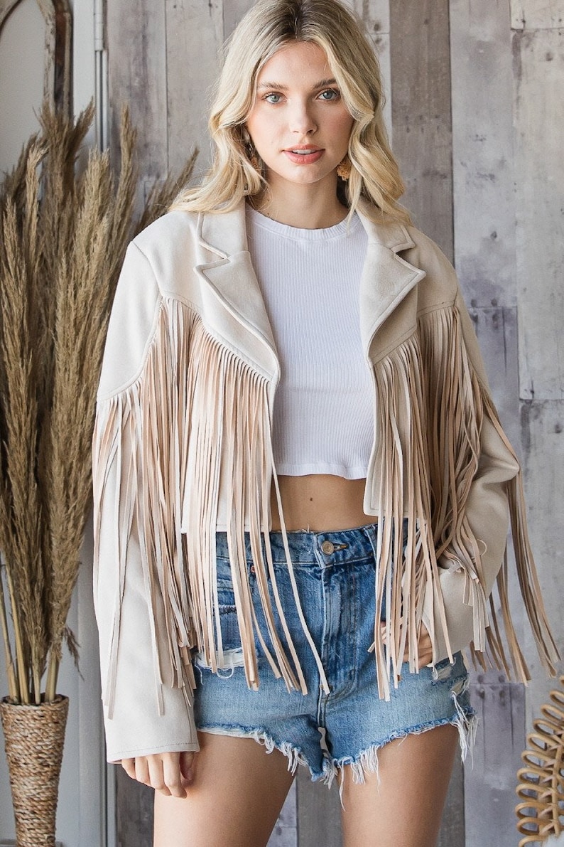 May include: Beige suede jacket with long fringe detailing. The jacket has a classic collar and is worn over a white crop top and denim shorts. The jacket's fringe adds a bohemian flair to the outfit.