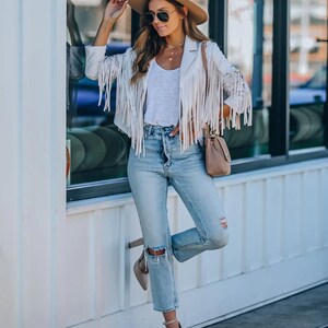 May include: A cream-colored fringed jacket, paired with light-wash ripped jeans and a white top. The outfit is accessorized with a tan fedora, sunglasses, and a small purse. The model is wearing high heels.