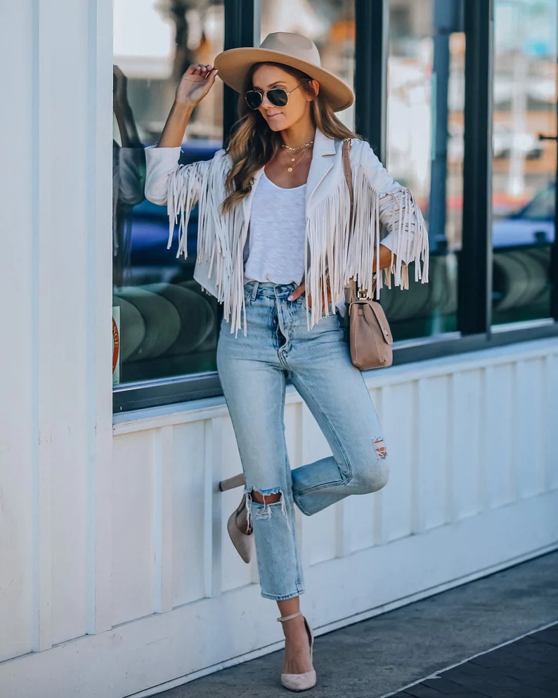 May include: A cream-colored fringed jacket, paired with light-wash ripped jeans and a white top. The outfit is accessorized with a tan fedora, sunglasses, and a small purse. The model is wearing high heels.