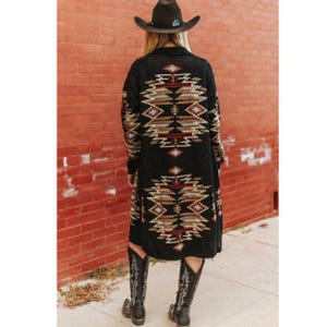 Könnte beinhalten: Langer schwarzer Cardigan mit einem geometrischen Muster im Südwesten in Braun-, Beige- und Rottönen. Der Cardigan wird mit einem braunen Cowboyhut und schwarzen Cowboy-Stiefeln getragen. Das Outfit befindet sich vor einer roten Backsteinmauer.